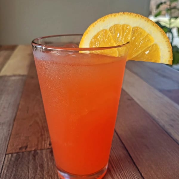 A vibrant orange cocktail with ice and an orange slice garnish sits on a wooden table.