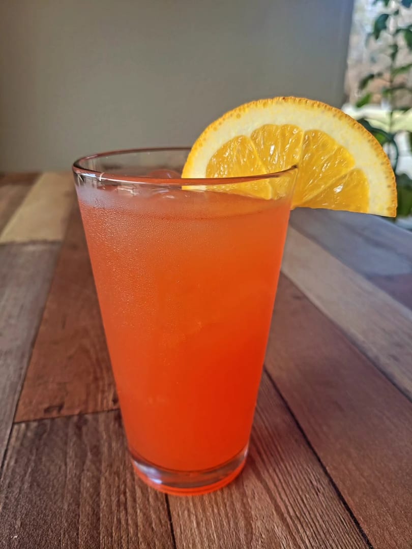 A vibrant orange cocktail with ice and an orange slice garnish sits on a wooden table.