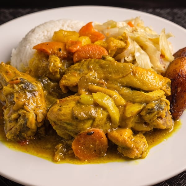 Curry chicken with rice, carrots, cabbage, and fried plantains on a white plate.