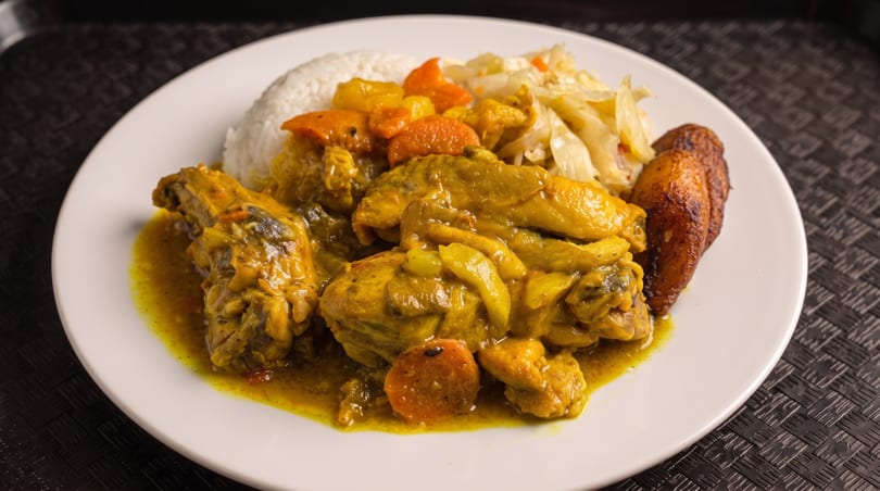 Curry chicken with rice, carrots, cabbage, and fried plantains on a white plate.