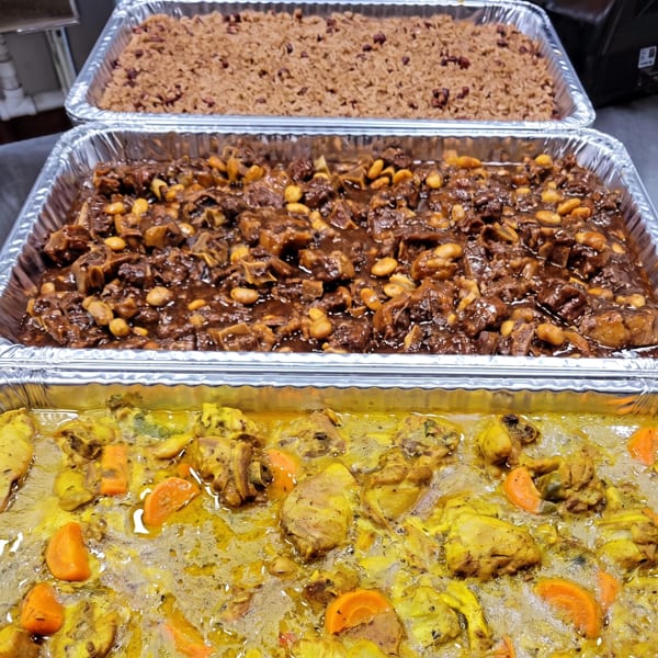 Three foil trays of Jamaican food: rice and peas, oxtail stew, and curry chicken with carrots.