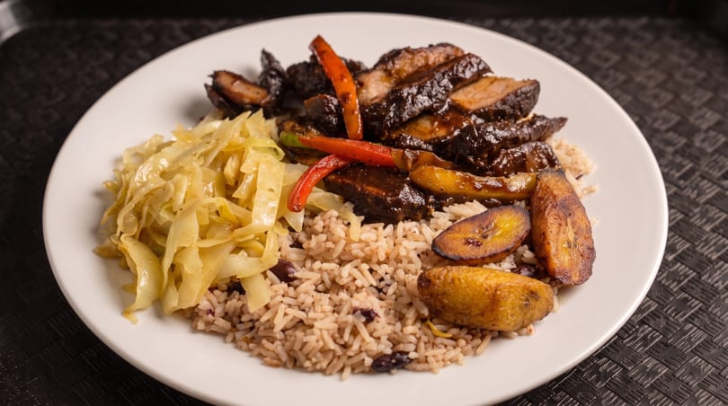 A plate of Jamaican jerk pork with rice and peas, steamed cabbage, and fried plantains.