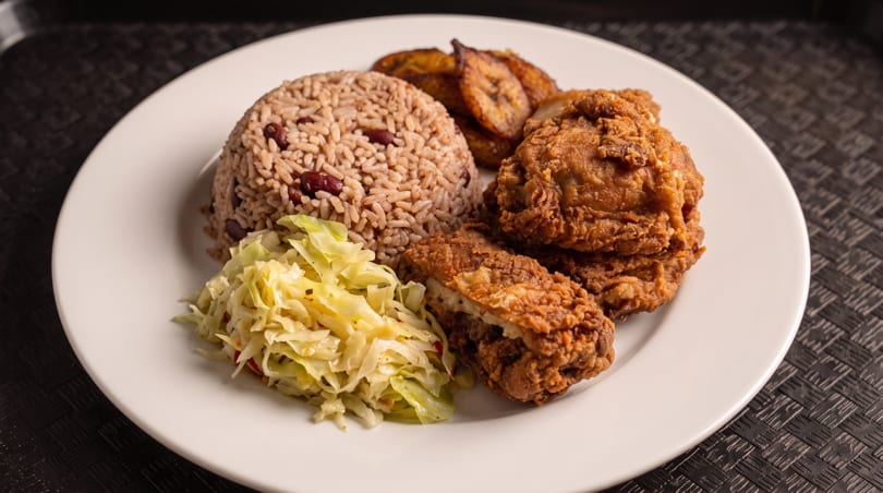 Fried chicken, rice and peas, plantains, and cabbage on a white plate.